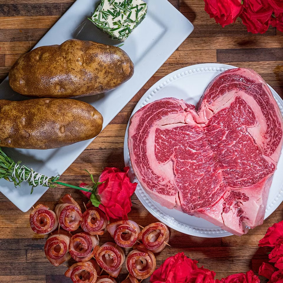 Heart-shaped ribeye steak with baked potatoes, bacon-wrapped appetizers, and roses for a Valentine’s dinner