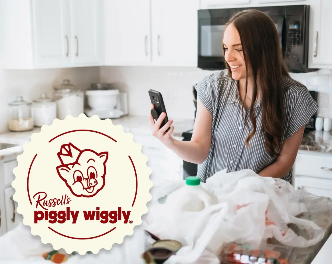 Woman smiling and using smartphone in a modern kitchen with grocery bags and Russell's Piggly Wiggly logo visible