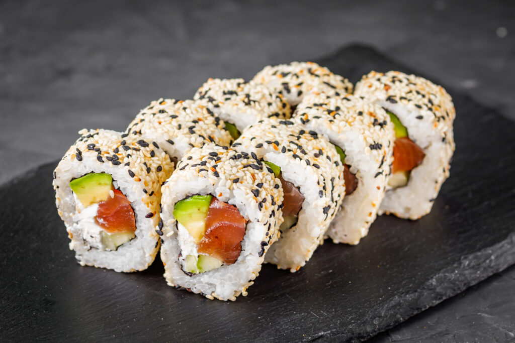 Close up of sesame coated sushi rolls with salmon avocado and cream cheese on a dark slate serving board
