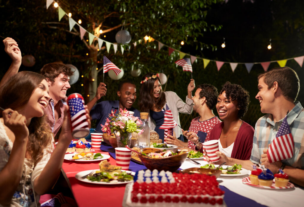 Group of friends celebrating the 4th of July at an outdoor backyard party with American flags, patriotic decorations, food, drinks, and festive lighting.